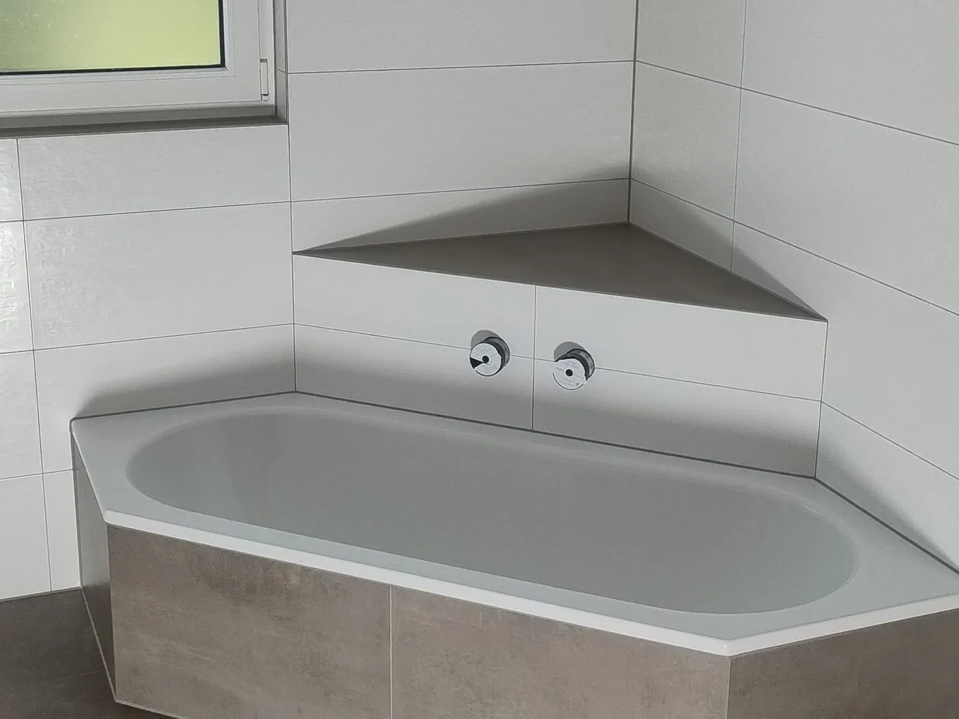 Eckbadewanne mit gefliestem Podest in Graubraun und weißen Wandfliesen mit Eckablage, Armaturen noch nicht montiert